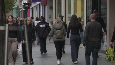 Two police officers walking on a crowded Exeter High Street. There are lots of people walking both towards and away from the camera, some of them holding carrier bags and backpacks.