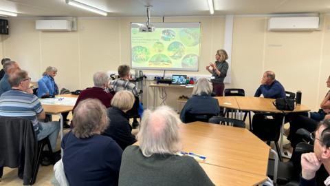 People at the public consultation sat in a room with various tables, listening to a speaker stood next to an overhead projector.