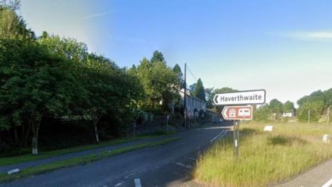 A road sign from the A590 into Haverthwaite. The road is lined by trees and there are a few houses visible in the distance.