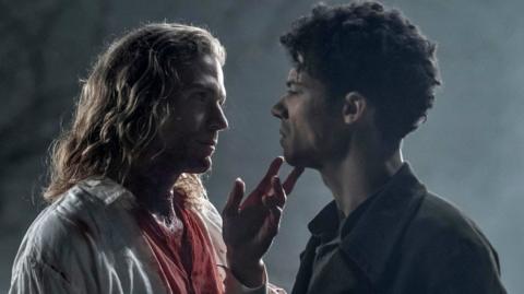 A still from Interview with the Vampire, showing two men, Lestat de Lioncourt (played by Sam Reid) and Louis de Pointe du Lac (played by Jacob Anderson) looking at each other while one touches the face of the other with bloodstains on their shirt. They are silhouetted against a grey-blue background.