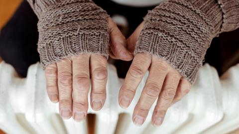 A pair of hands wearing fingerless gloves are being warned on a white radiator.