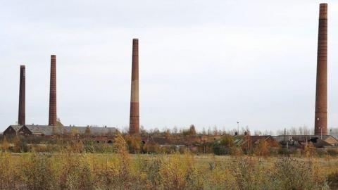 Four chimneys at a former brickworks, near Bedford. They stand tall against a grey sky. There are a few brick buildings between the chimneys and scrubland in the foreground.