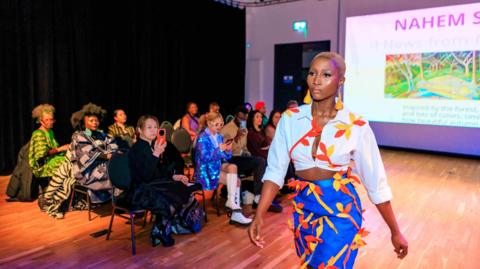 A woman wearing a white cropped shirt and a blue skirt walking down a cat walk. She is wearing orange dangly earrings and her outfit has a yellow and orange leaf pattern on. People in the audience are sat on black chairs taking pictures of her outfit at a fashion show.