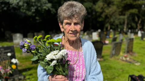 A woman stands in front of a grave holding flowers