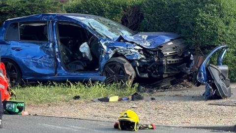 A side view of a crumpled up blue car on a pavement and half buried in a thick hedge. Its bonnet has buckled, its windscreen is a haze of cracks and its driver's side door has been removed and is resting on a driveway in front of the bonnet. There is a yellow emergency services helmet resting on the road in front of it, beside some gloves.
