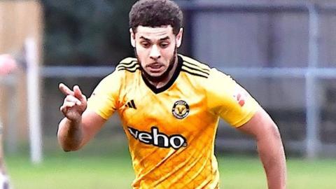 Man in bright yellow football shirt with black detailing he has dark brown hair.