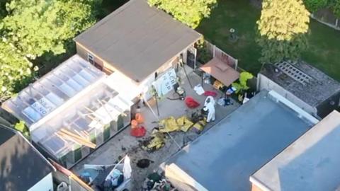 A drone image of the backyard at Save A Paw centre. Forensic officers wearing white jumpsuits are examining the scene, which is messy with various bags, mud and yellow sheets laid across it. There is a large grey outbuilding with a flat roof. Next to it is a smaller building with transparent roof.