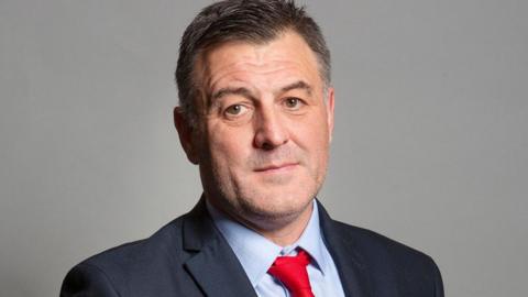 The MP has greying dark, a light blue shirt, red tie and and dark blue  jacket.