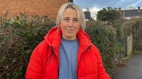 A woman with light blonde hair tied back, wearing a blue sweater and a bright red puffer jacket with black accents