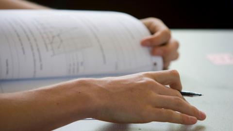 A student opens an exam paper with pen in hand. Only their hands are visible beyond the pen and paper.