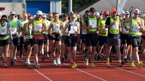 Lines of participants wearing neon yellow vests with entry numbers on them power walking on the NSC track at the start of the walk.