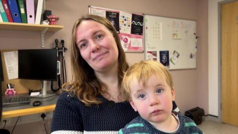 Ros and her son Eddie in a GP's room, both wearing jumpers and both with blonde/light brown hair. They are both also looking at the camera. 