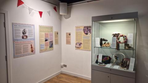 A glass cabinet with artefacts, including shoes and jewellery, highlighting Polish culture. It is standing in a museum with posters and information boards on a crisp white wall.