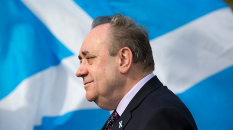 Alex Salmond is side-on to the camera with a Saltire flag behind him