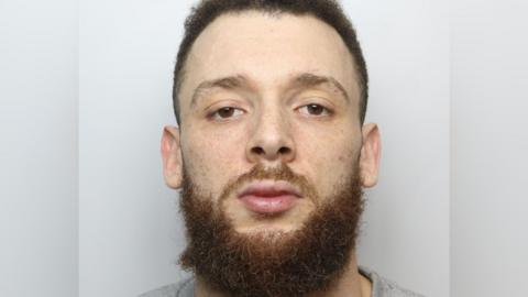 A mugshot of a man with brown eyes, short brown hair and a long orange beard. He is wearing a grey shirt.