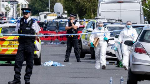 Police and forensics on Hart Street Southport, Merseyside, after stabbings of children at a dance class. There is police tape cordoning off the road, several cars and two people in white suits 