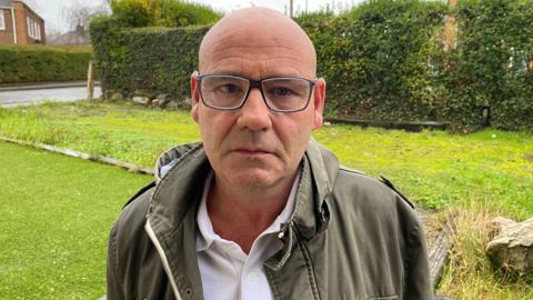 Barry Froggatt in a picture. He is standing on a front drive looking at the camera. He is making a serious face. He is a bald man wearing glasses, a white polo top and a green jacket. 