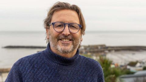 Mitch Tonks stands in front of Lyme Regis harbour on a gloomy day. He is smiling and wearing a navy blue turtle neck jumper.