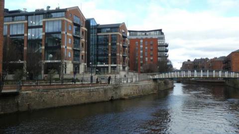 The River Aire in Leeds city centre, with the David Oluwale Bridge in the distance. Flats and offices are on the other side of the water. The sky is blue with some cloud cover. 