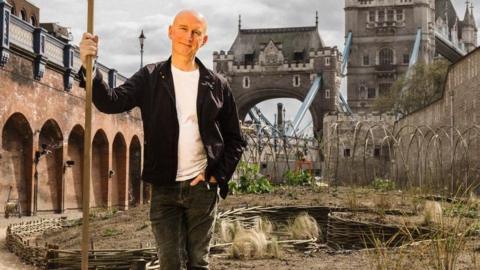 Landscape gardener and horticulturalist Nigel Dunnett wearing black jacket and trousers and white t-shirt, holding a stick with the Tower of London behind. Dunnett designed the wildflower project for Superbloom in the Tower of London moat, (date).