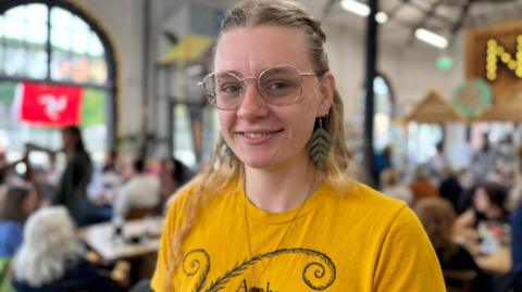 Celia Bales has long blonde hair, half tied up. She is smiling, wearing glasses and a yellow t-shirt. She is standing in a busy cafe with high ceilings.