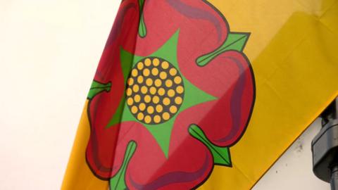 An image of a Lancashire flag flying. The flag is yellow with the Lancashire Red rose on it.