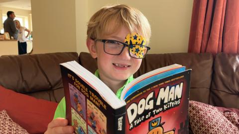 Harry is sitting on a couch reading a book called Dog Man. He is smiling at the camera wearing glasses and a patch over one eye with rockets and stars on it. 