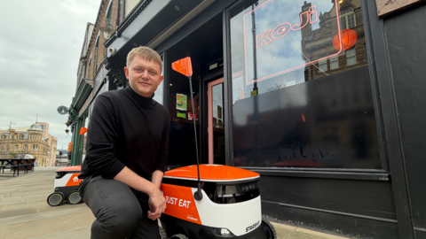 Peter Richardson, from Koji, kneels next to the white-and-orange branded robotic vehicle, which has a black pole with an orange flag on top. He is outside the restaurant on High Street West, and in the background is another robotic vehicle.