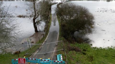 A 'road closed ahead' sign and green barriers block a road flooded with high waters on either side of it near to Harbridge in Hampshire.
