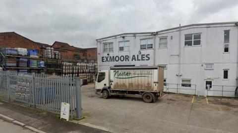 A photo of the factory in Wiviliscombe from the outside, a white two storey building with barrels of beer stacked up outside and a truck parked in front. 