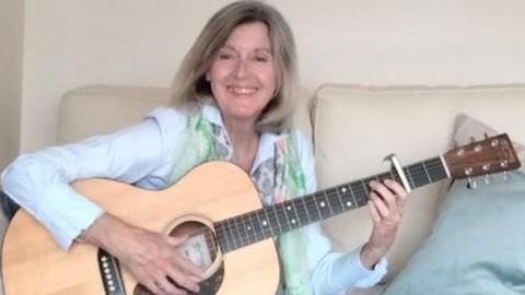 Roe Dickson smiling as she sits on a sofa holding her guitar with her fingers on the fret board