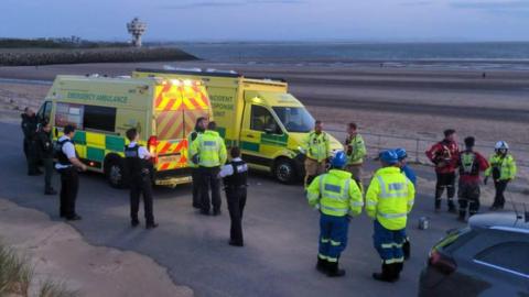 More than a dozen emergency service workers in high-visibility jackets and police uniforms wait near two ambulances. They are next to the sands of Crosby beach around sunset with the sea in the background.