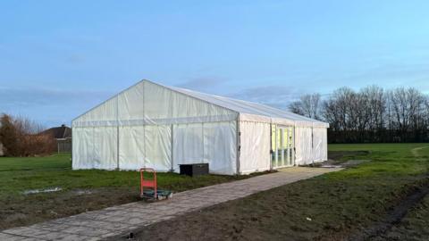 A white gazebo-style marquee in the middle of a muddy grass field. The marquee has glass French doors on its right hand side. There is a light-coloured paved path leading up to the French doors. In the background, a blue sky and leafless trees. 