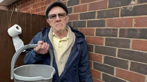 Seventy-nine-year-old John Grimley stands in front of a wall and a garden gate holding a grey bucket.  He is wearing a navy anorak and a grey peaked cap. Toilet roll is hanging on a hook behind him