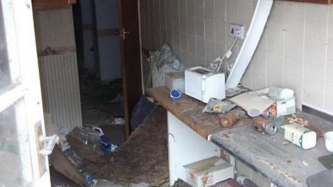 The dirty kitchen of the home. There is litter everywhere, a carpet pulled up on the floor.