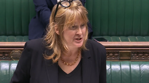 A woman with long fair hair stands at the Commons despatch box with green benches behind her. She is wearing a black suit and top, and a gold necklace.