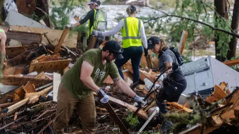 A man and woman in the foreground wearing blue caps, short-sleeved shirts and dark trousers remove piles of debris, two other women walking among wooden debris in the background
