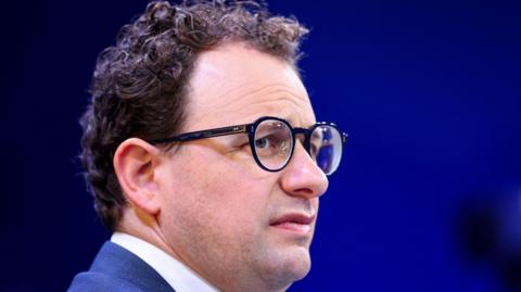 Anthropic CEO Dario Amodei on a stage, furrowing his brow behind round, black-rimmed eye classes.