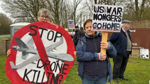 Two women stand in front of other protesters near an RAF Fairford entrance sign. They are holding signs, one is round and in the format of a giant stop sign in red. It features a painted military drone and the words "Stop drone killing". The other sign simply states "US war mongers go home" in black capital letters.