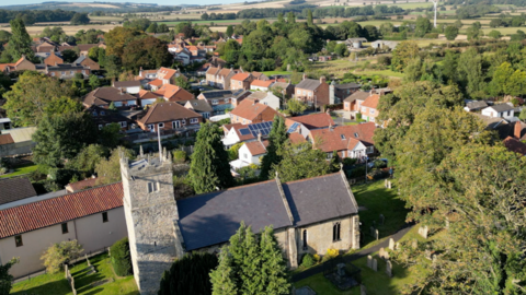 A church and several houses in Shiptonthorpe can be seen from above. The sun has cast a shadow on the church and highlights different hues of green within nearby trees.