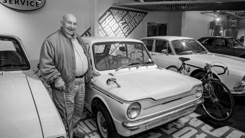 A smiling Brian Pollard, wearing a shirt and jacket, holds a walking stick. He is standing next to a Hillman Imp car, a vehicle he worked on during his time at Humber. Other cars can be seen in the background of Coventry Transport Museum