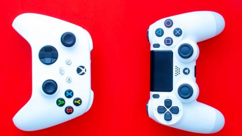 An Xbox (left) and PlayStation controller (right) side by side on a deep red background viewed from above. Both are the white versions of the device and the shoulder buttons of each are facing each other. The button layouts are standard.