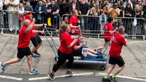 A mixed gender team of runners wearing red tops and black running shorts push a metal trolley between them, with one person sat in the middle. In the background a crowd of people behind a metal barrier can be seen cheering them on.