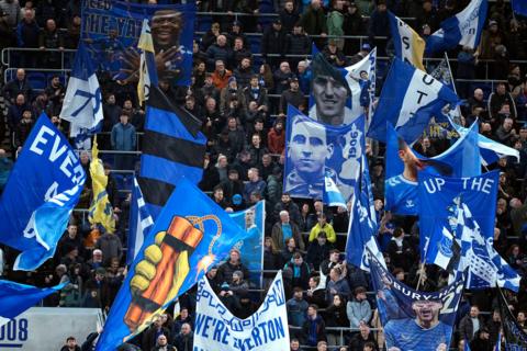 A packed football stadium full of Everton fans, many of them waving their club's blue flags.