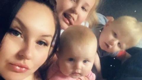A young mum pictured alongside three young blonde-haired children. The mum has dark hair.