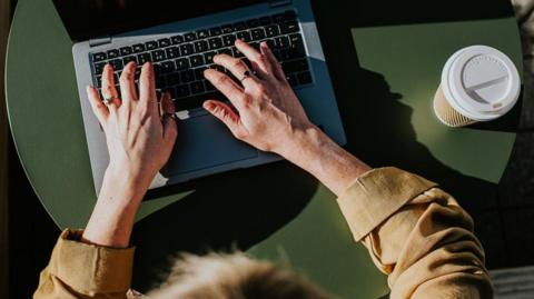 A woman sits at a laptop with her hands over the keyboard and a disposable cup and lid beside her.