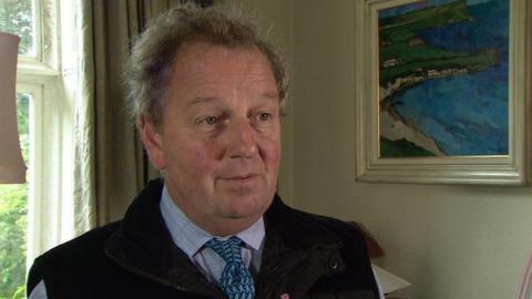 Danny Kinahan, sits in front of a window, with a painting of the sea to his left. He is wearing a shirt and tie