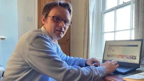 A man, wearing a grey jumper over a white shirt, is sitting at a desk. He has turned to the side to smile at the camera. There is a laptop on the desk and his hands are resting on the keyboard. He has short brown hair and is wearing rectangular framed glasses. 