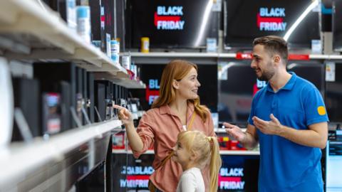 Salesman speaks to mother in a shop surrounded by TV's that say Black Friday. She is joined by her young blonde daughter.