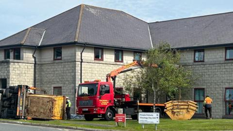A building in the background, you can see a red truck with a crane on a field in front, a skip truck has tipped over on its side.